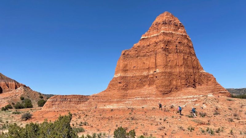 Cathedral Peak, a tall layered red sandstone peak under blue sky with small hikers at its base