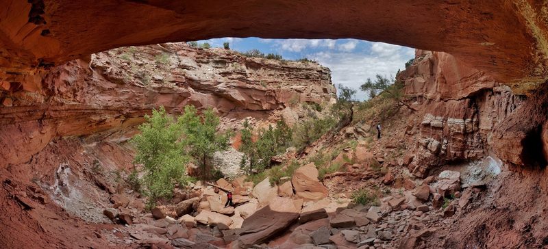 Panorama from beneath a red sandstone overhang looking out into Red Canyon, with cottonwoods, boulders, and two small hikers below